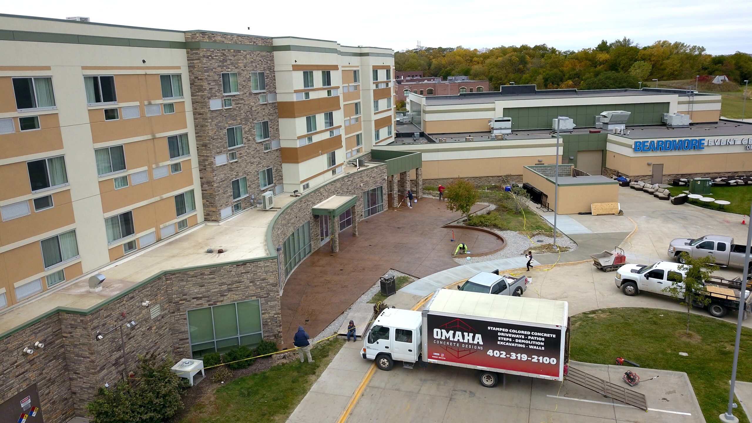 Aerial view of Beardmore Event Center with a hotel, parked vehicles, workers on site, and a moving truck labeled “OMAHA” near the entrance. Trees and additional buildings are in the background.