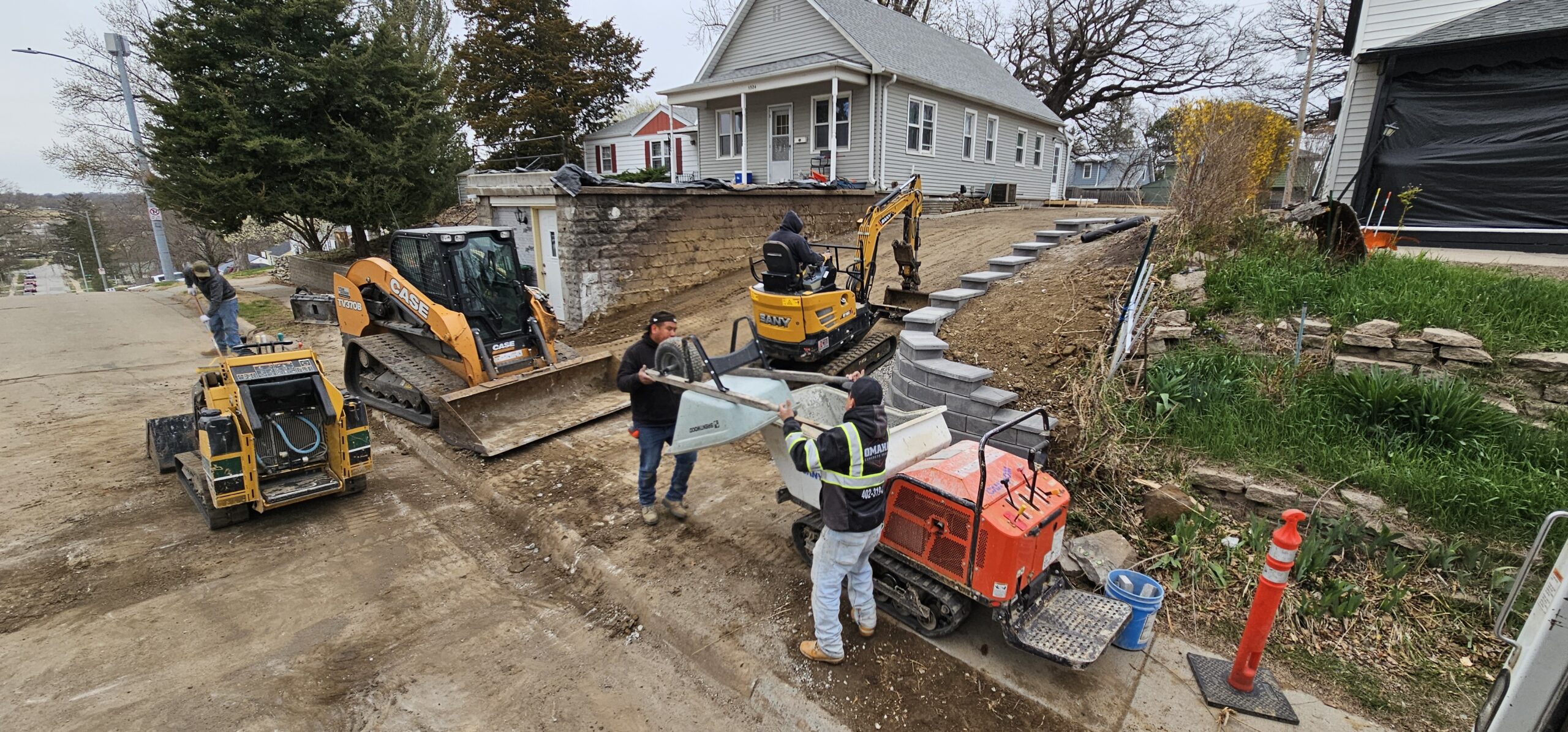 Workers use construction equipment and tools to perform landscaping and grading work on a residential property next to a street, while a concrete contractor prepares the site for paving.
