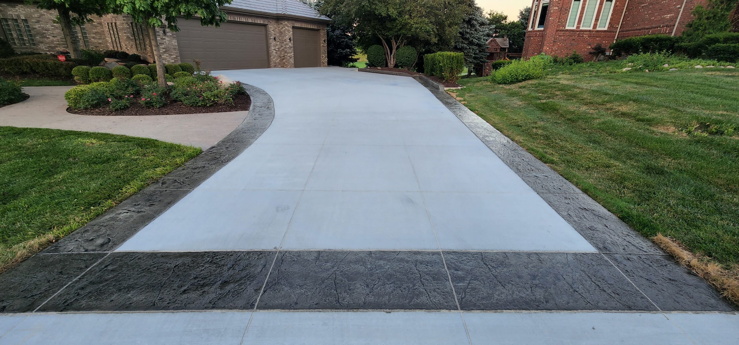 A clean, newly paved concrete driveway with decorative dark borders leads up to a two-car garage beside a manicured lawn, highlighting the home's elegant concrete driveways and walkways and landscaped garden.