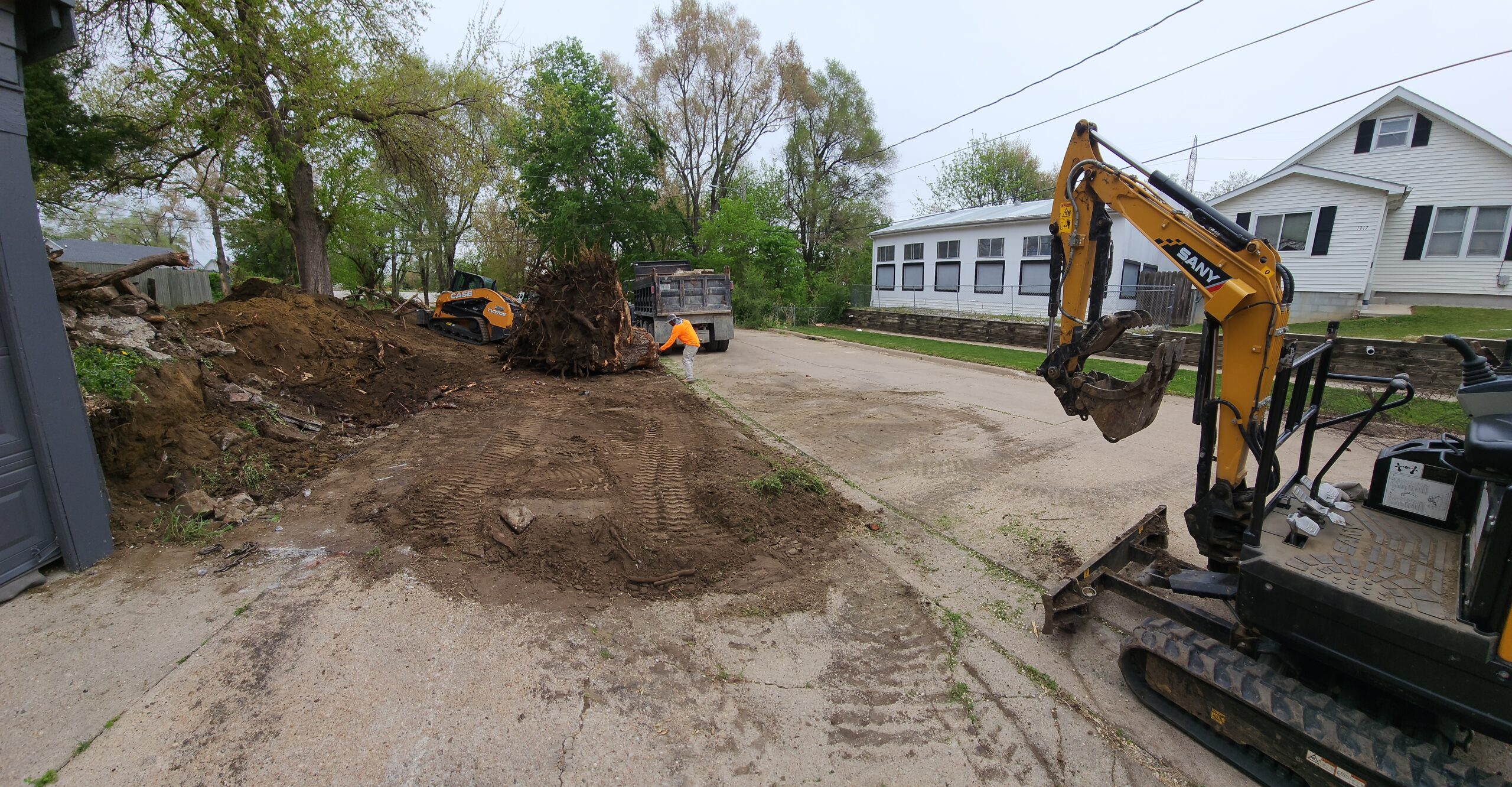 A small excavator and debris pile are on a residential street, where concrete repair and restoration work is underway, with a dump truck and dirt visible near houses and trees.