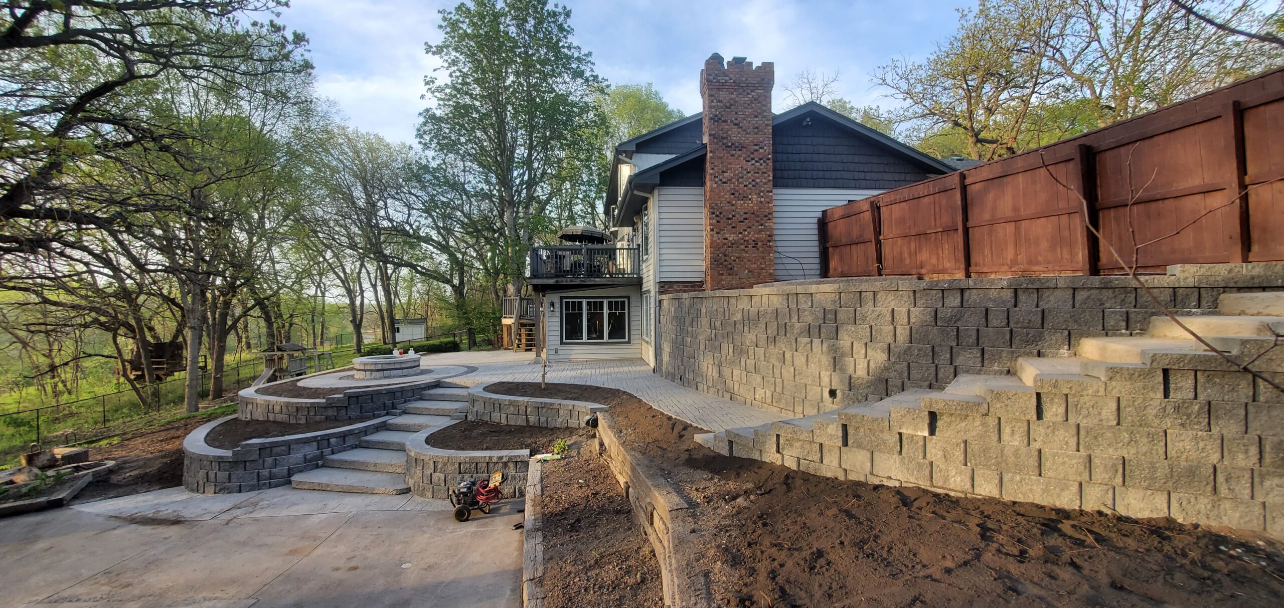 A backyard with retaining walls, a partially finished patio, fire pit area, and a two-story house with a deck and chimney, surrounded by trees.