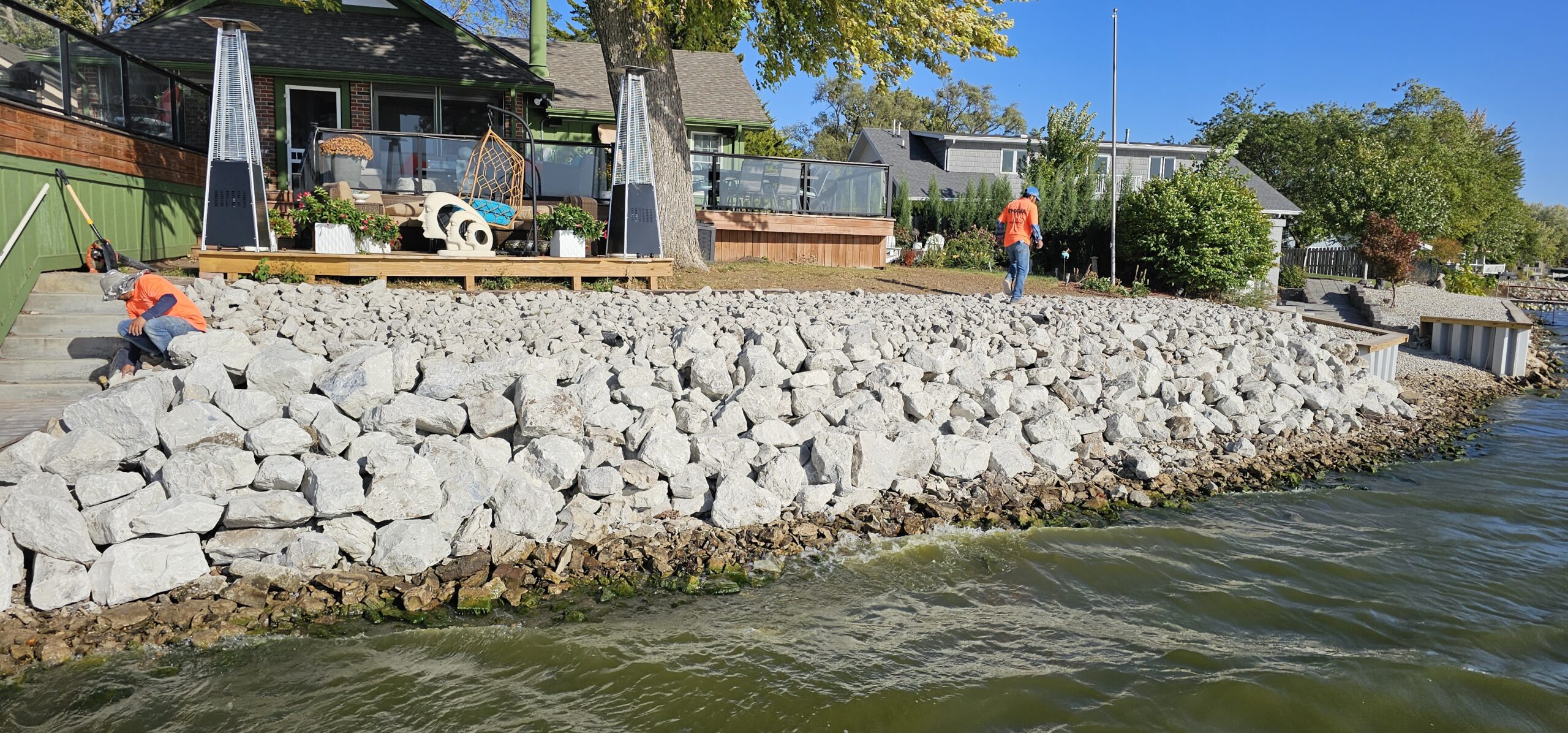 Two workers in orange shirts are placing large rocks and working on concrete retaining walls along a shoreline in front of lakeside houses under a clear blue sky, enhancing the area's hardscaping.