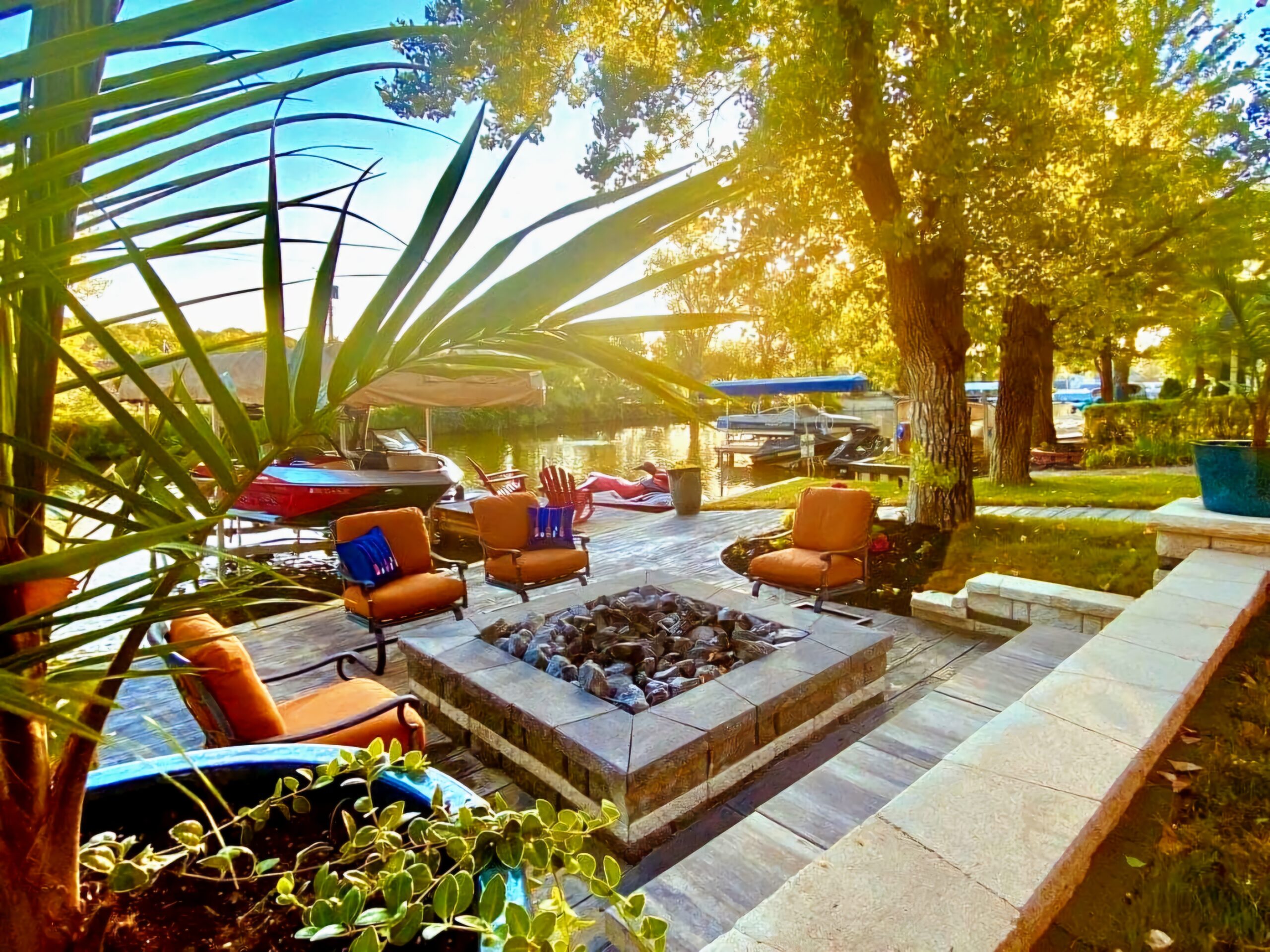 A lakeside patio with cushioned chairs around a fire pit, boats docked nearby, and trees providing shade in the sunlight.