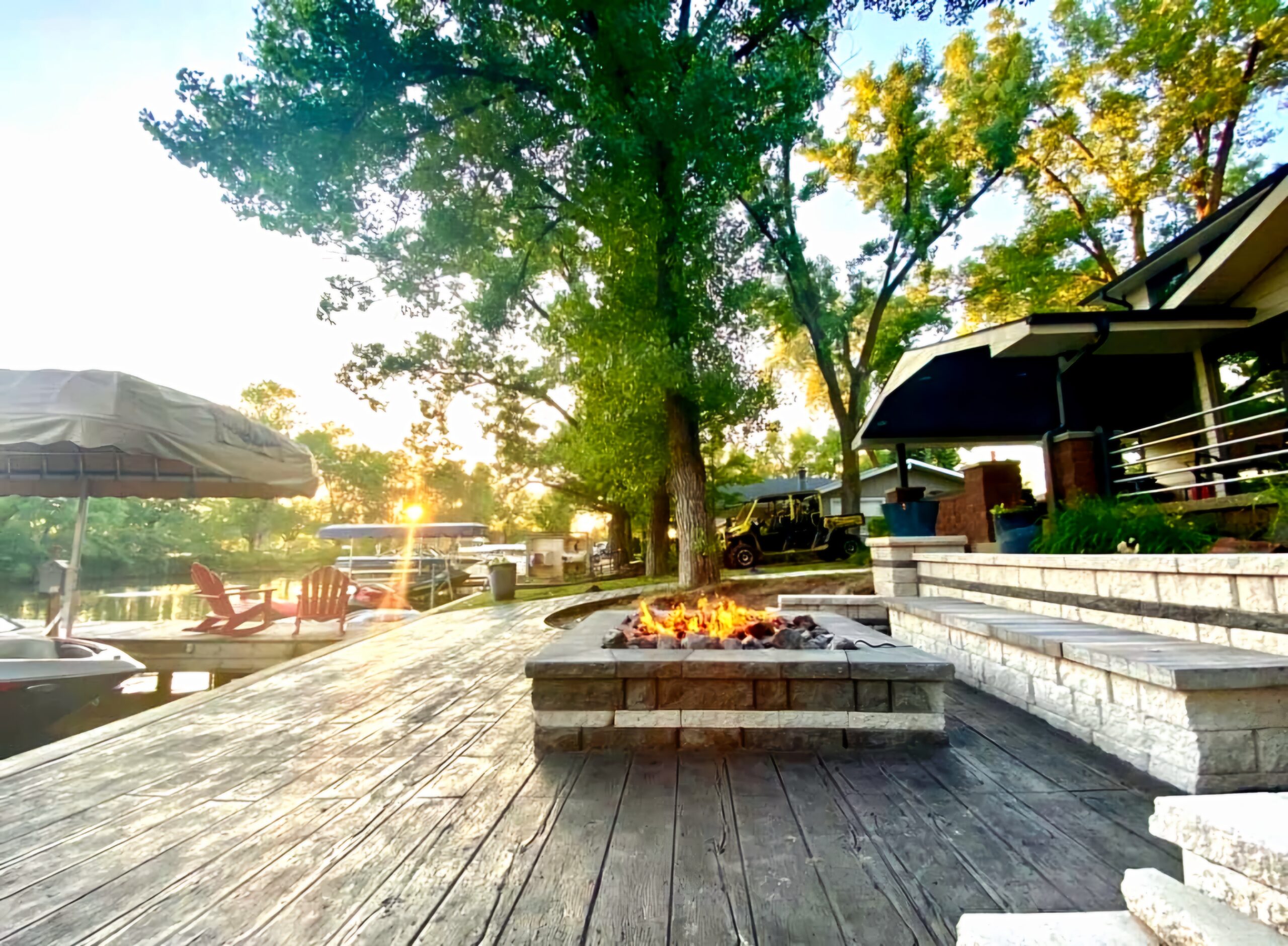 A backyard patio with a fire pit, wooden deck, and seating overlooks a calm lake with boats and trees at sunset.