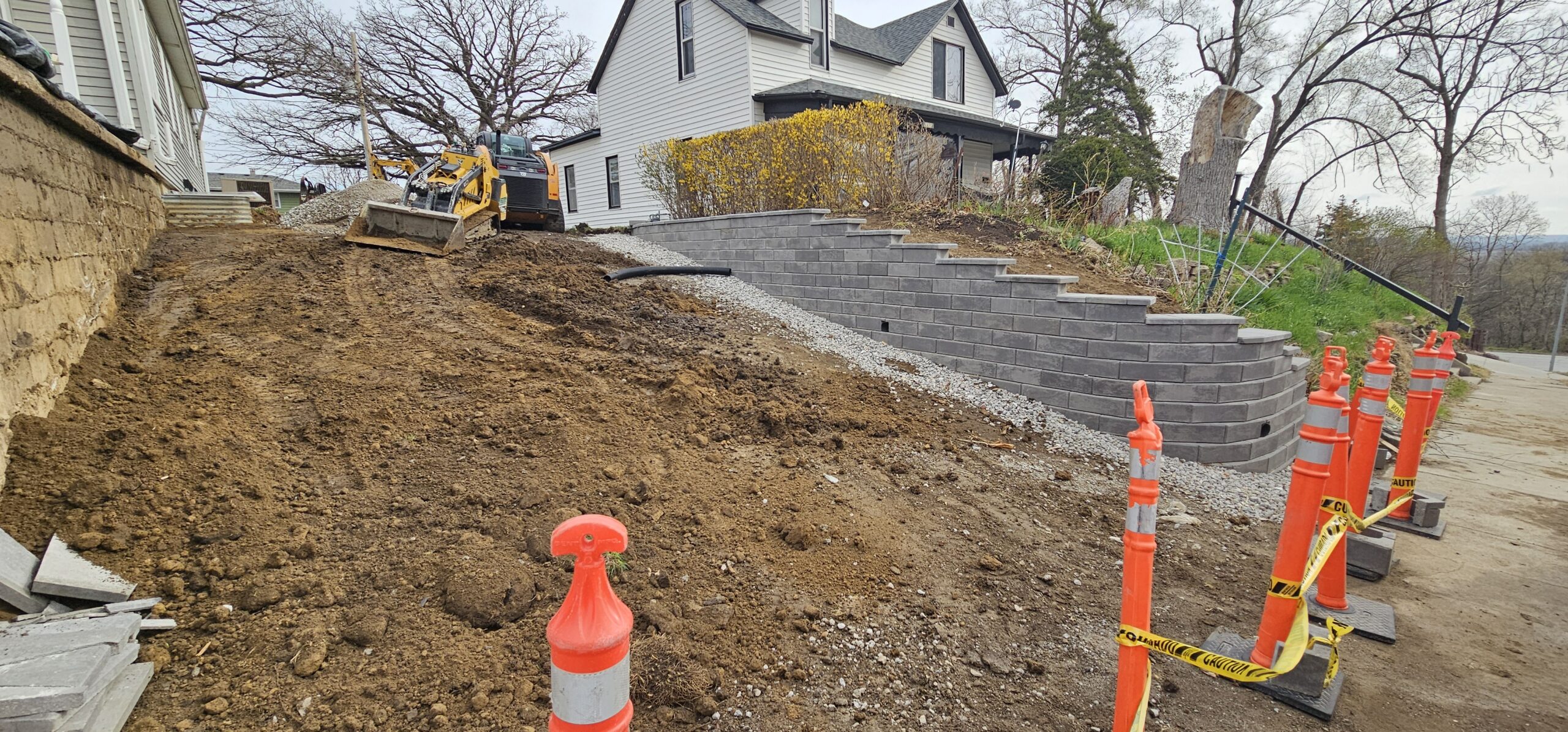 A construction site with a skid steer grading soil next to a house, a new retaining wall, and orange safety cones marking the work area.