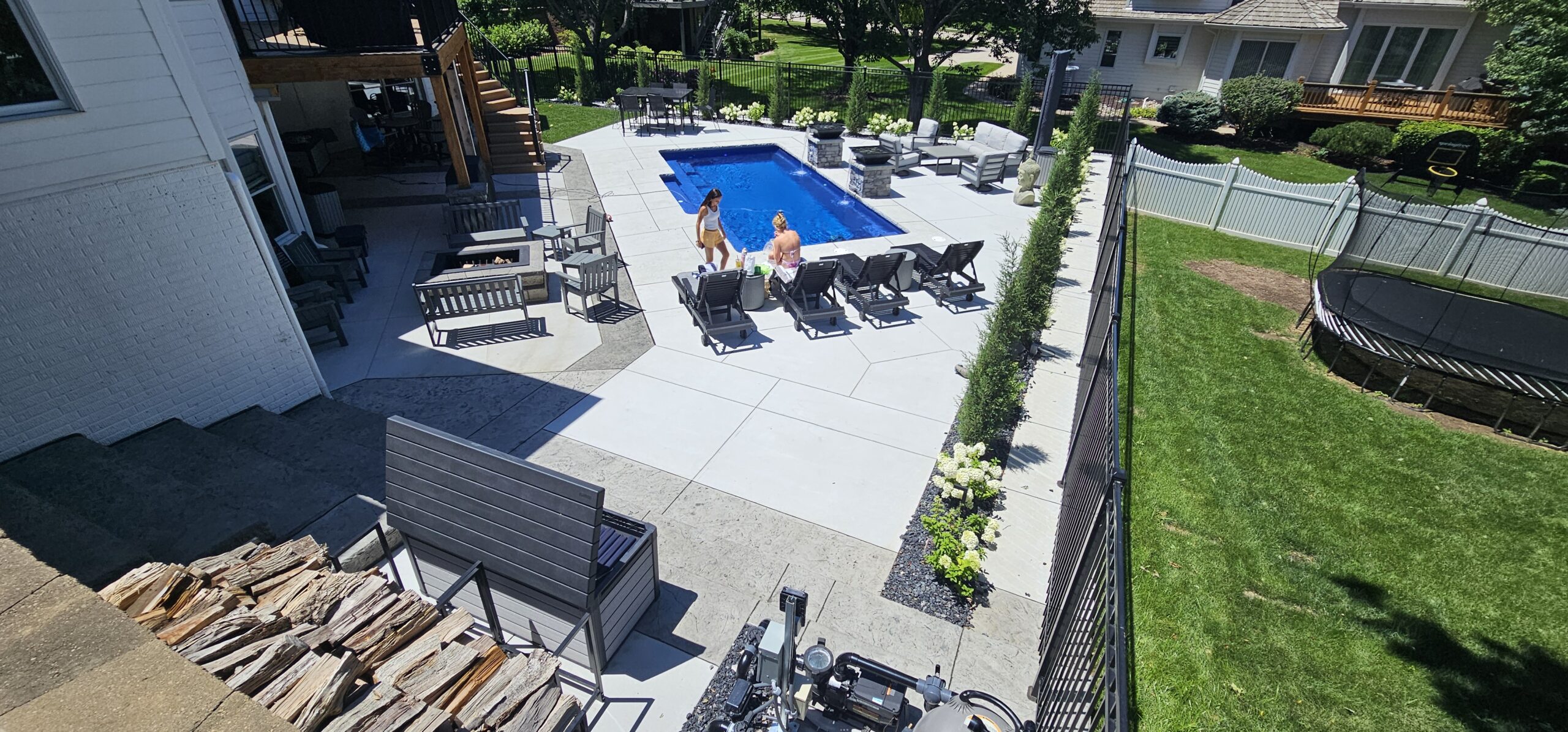 A backyard with a rectangular pool, lounge chairs, and people standing near the pool; surrounded by patio furniture, greenery, and a fenced grass area with a trampoline.