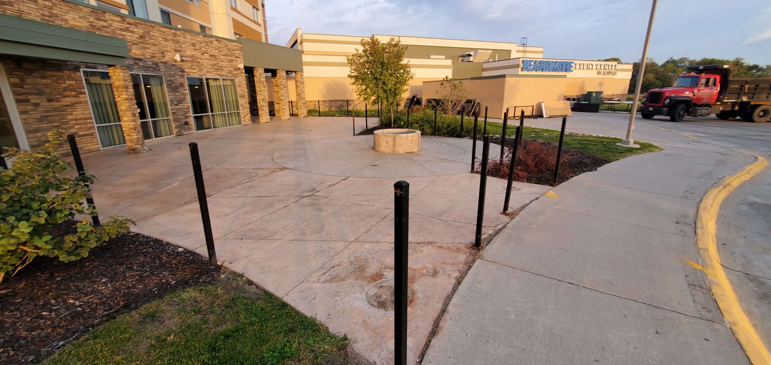 A circular concrete patio with a central planter is surrounded by black metal posts near a building entrance and a parking lot, under early evening sunlight.